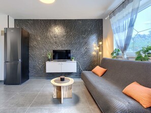 A modern living room with a marble wall, a flat-screen TV, a built-in white cabinet, a grey sofa with orange cushions, a wooden coffee table, and a refrigerator.