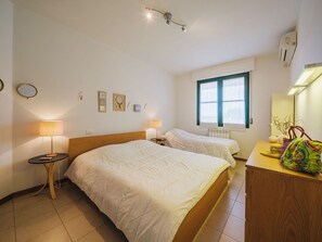 Hotellihuone, jossa on kaksi sänkyä, yöpöytä lampulla, lipasto, ikkuna, jossa on sälekaihtimet, ja seinäkoristeita.