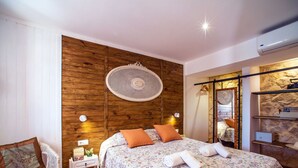 Une chambre avec un mur en bois, un lit recouvert d’une literie à motifs floraux, une chaise en osier et un ventilateur suspendu au plafond.