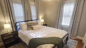 A neatly arranged bedroom with a large bed, two bedside tables, a nightstand, and a floor lamp. - Classy Copacetic Cottage-King bd, Clean, close to Downtown, Furman, Swamp Rabbit (Greenville)