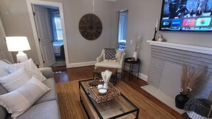 A living room with a sofa, coffee table, TV, fireplace, and a clock on the wall. - Classy Copacetic Cottage-King bd, Clean, close to Downtown, Furman, Swamp Rabbit (Greenville)