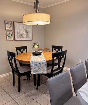A dining area with a round table, six chairs, a floral table runner, a vase with flowers, and a wall-mounted light fixture.