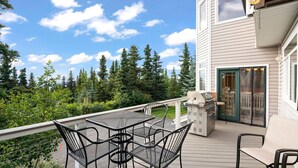 A deck with a grill, outdoor seating, and a view of a forested area. - Hillside Haven (Anchorage)