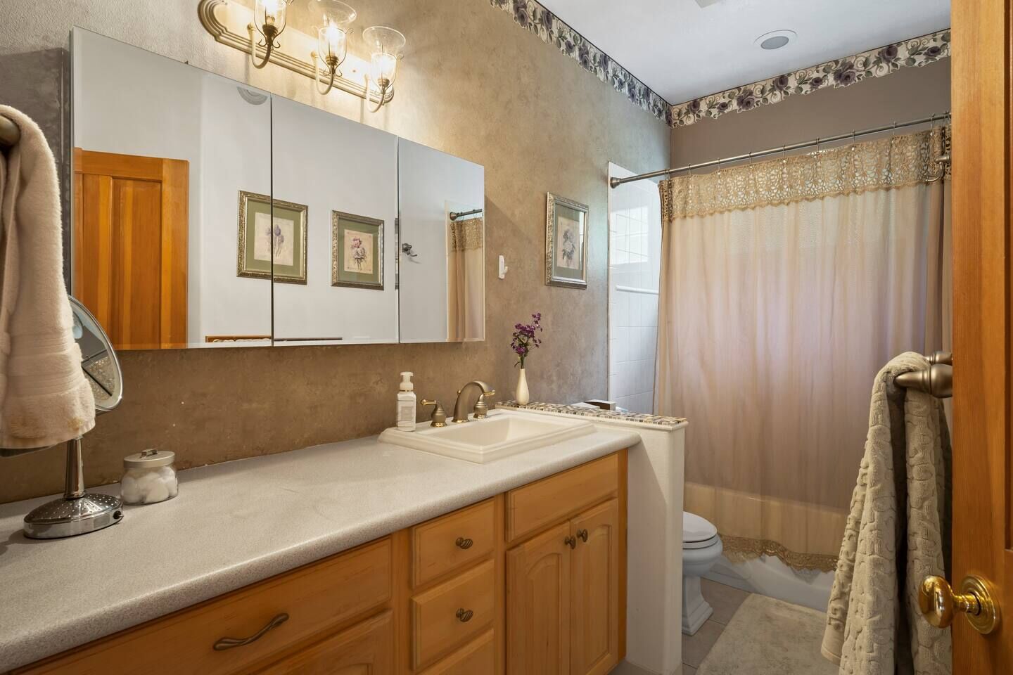 A bathroom with a vanity, sink, and mirror. There are framed pictures on the wall, a vase with flowers, and a shower curtain.