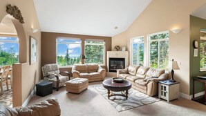A spacious living room with a fireplace, large windows, and a view of the outdoors. - Hillside Haven (Anchorage)