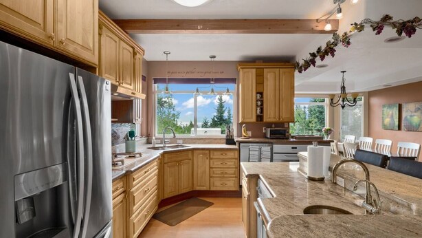 A modern kitchen with wooden cabinets, stainless steel appliances, granite countertops, and a large window offering a view of trees. - Hillside Haven (Anchorage)
