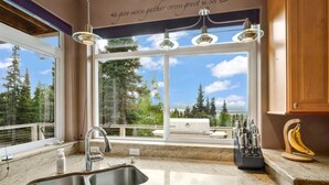 A kitchen with a large window, a stainless steel sink, and a countertop with a knife block and a banana holder. - Hillside Haven (Anchorage)