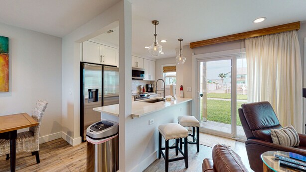 A modern kitchen with stainless steel appliances, a dining area with a wooden table and wicker chairs, and a living room with a leather sofa and a glass coffee table. - First Floor Condo, Faces Shared Pool, Golf Cart Access (Port Aransas)
