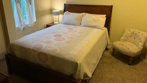 Un lit bien fait, avec une couvre-lit à motifs, une lampe de chevet, une chaise et une fenêtre avec des rideaux.