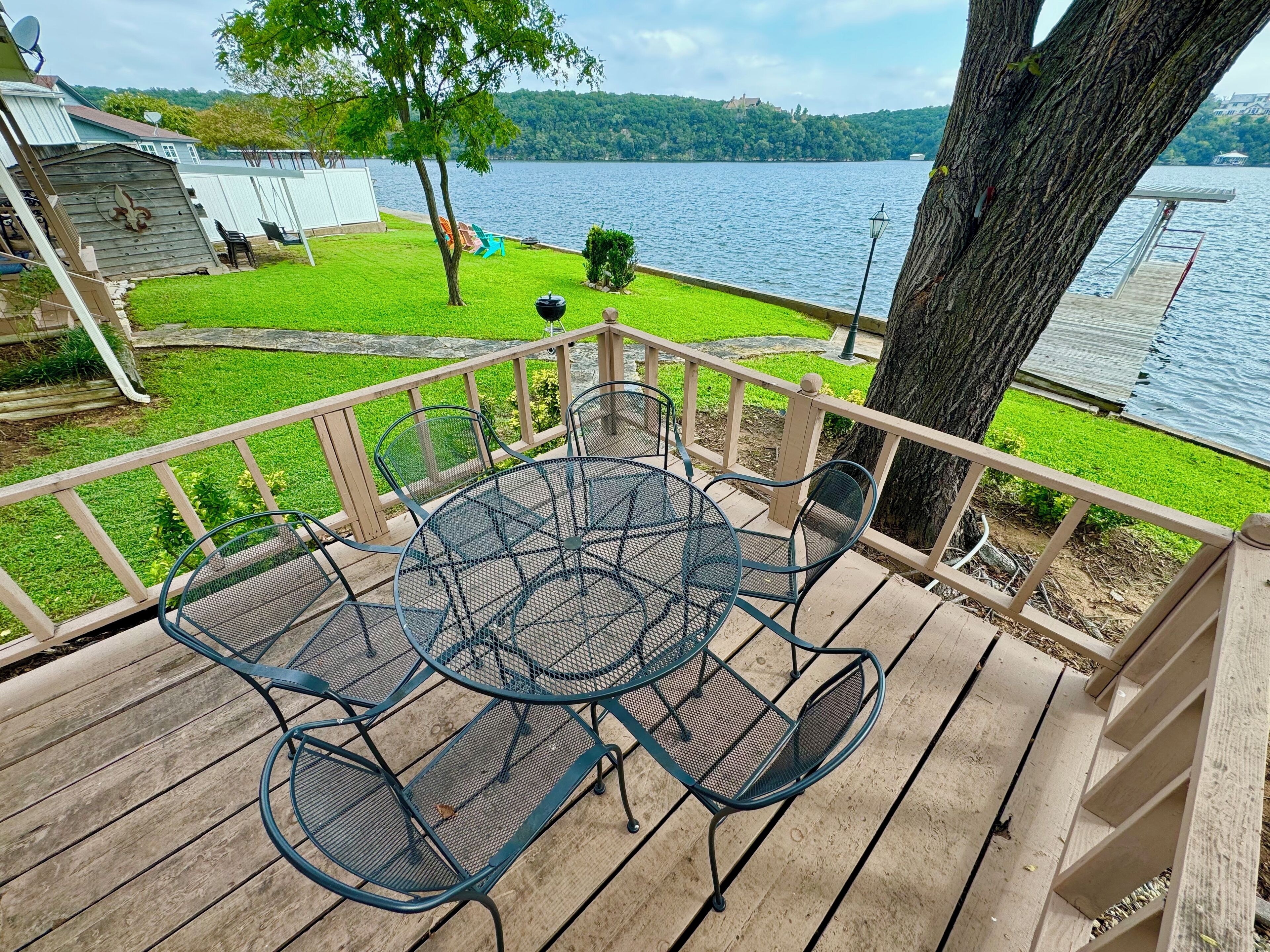 A wooden deck with a metal table and chairs, overlooking a lake with a grassy area and a building in the background.