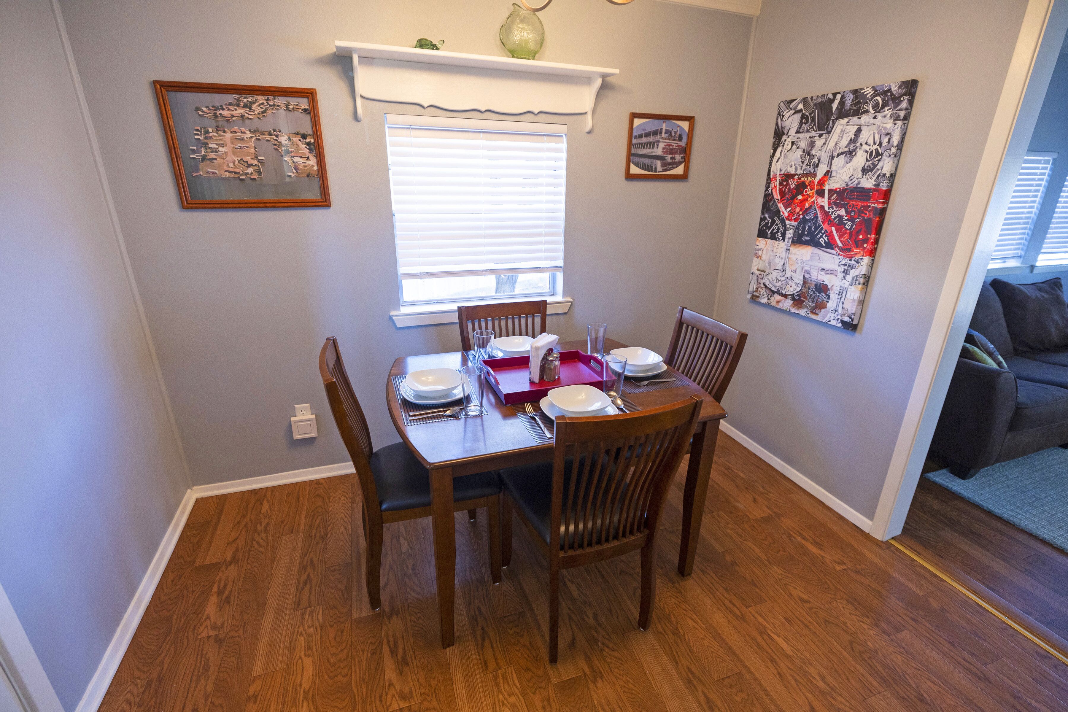 Un comedor con una mesa preparada para cuatro personas, una ventana con persianas y un cuadro en la pared.