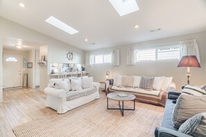 A spacious living room with a white sofa, a patterned sofa, and a coffee table. There is a kitchen area in the background with a window and curtains. - Harris Beach Haven | Hot Tub, Theatre & Fire Pit (Brookings)