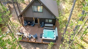 A cabin with a hot tub, deck chairs, and a covered seating area surrounded by trees.