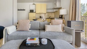 Un salon moderne avec un canapé gris, une table basse sur laquelle sont posés des livres et des sous-verres, un téléviseur sur un meuble, et une cuisine en arrière-plan.