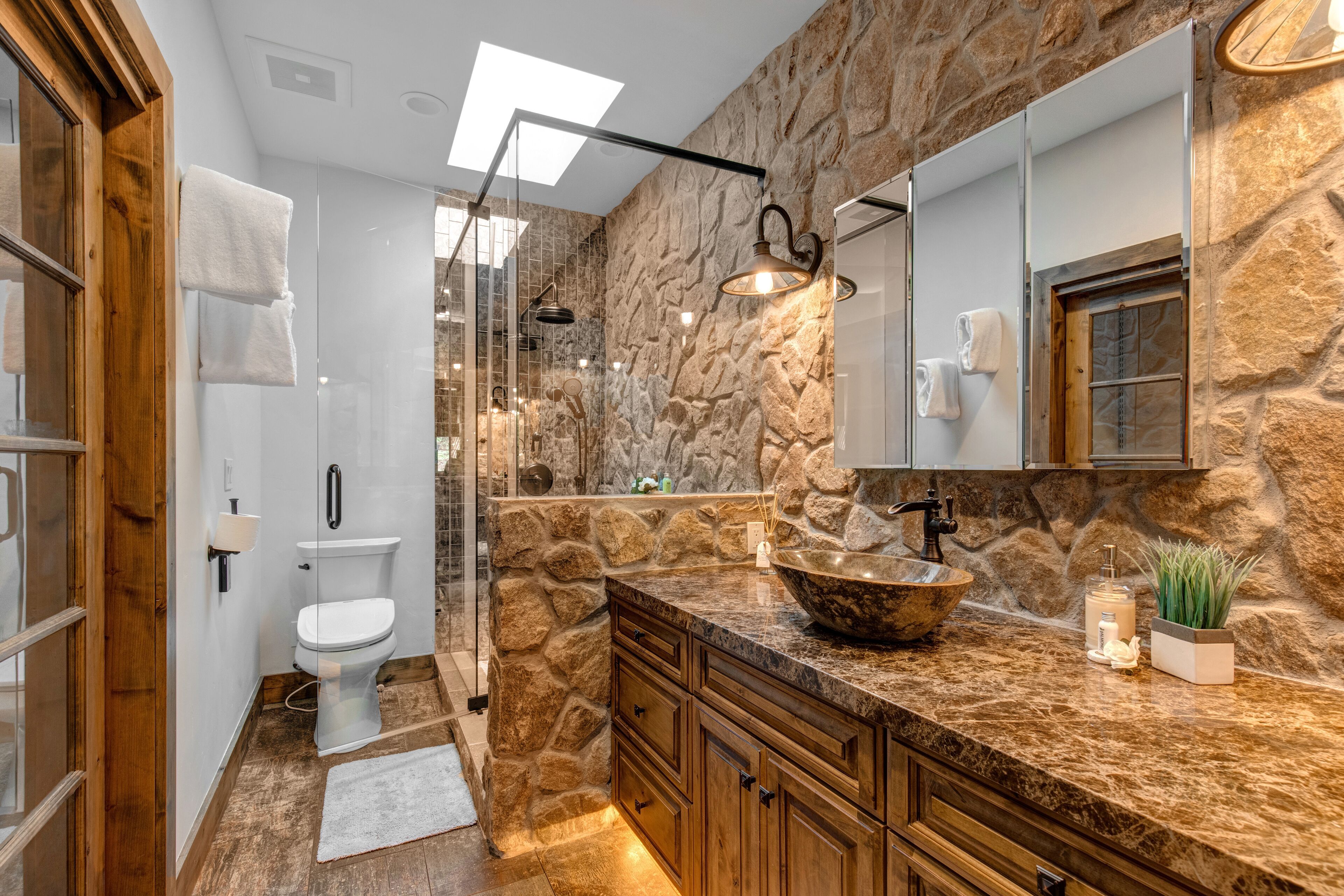A bathroom with a stone wall, a wooden vanity, a glass shower enclosure, and a large mirror.