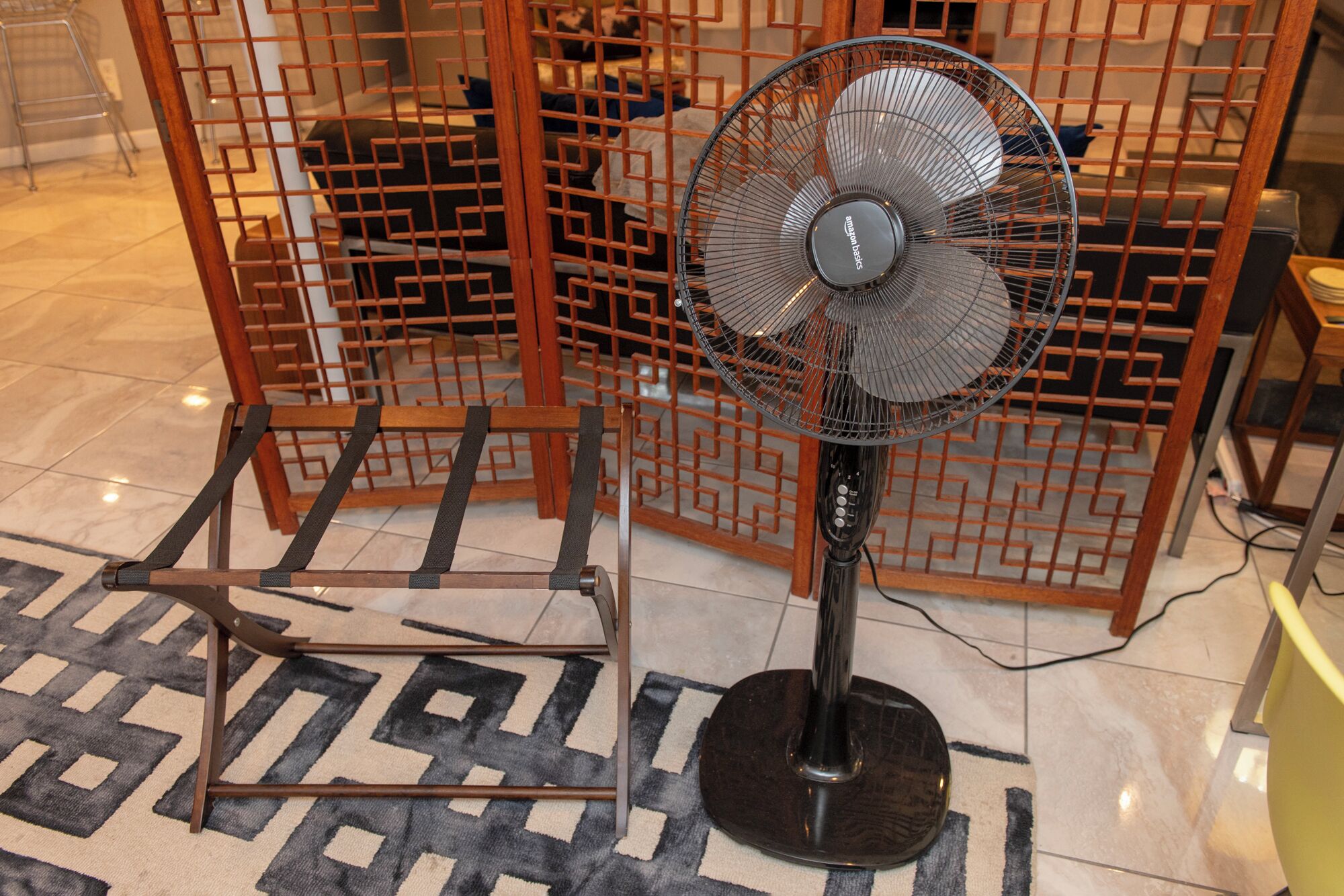 Een staande ventilator met metalen rooster en een zwart voetstuk, geplaatst op een vloerkleed met een patroon in een kamer met betegelde vloer en een metalen tussenscherm met geometrische patronen op de achtergrond.
