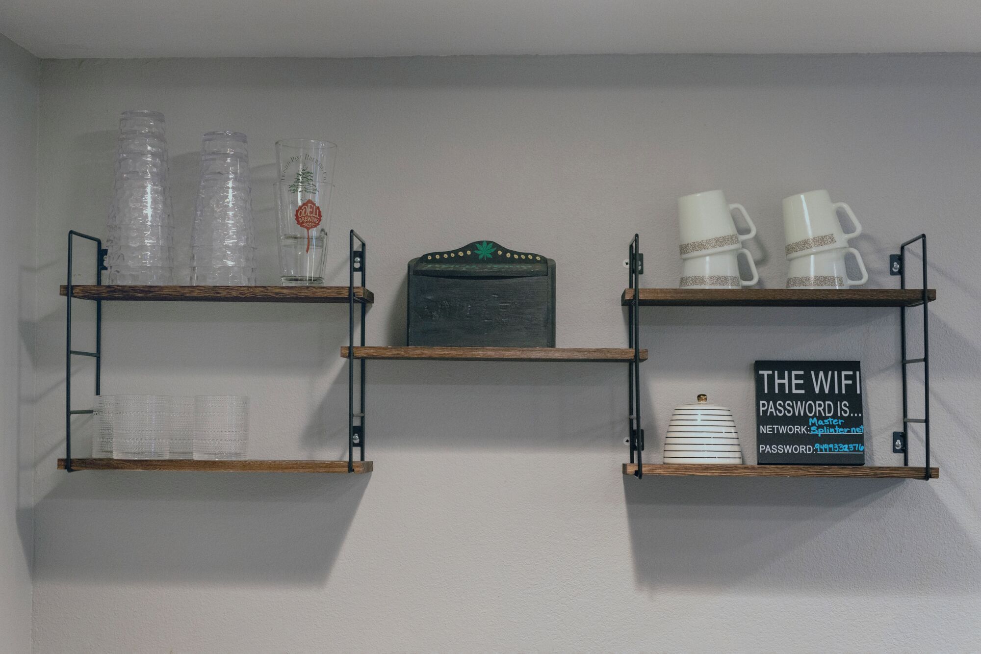 Tres repisas de madera con varios objetos: a la izquierda, botellas de plástico y un vaso con un logotipo; en el centro, un letrero con un logotipo; a la derecha, tazas blancas y un letrero con la contraseña de WiFi.
