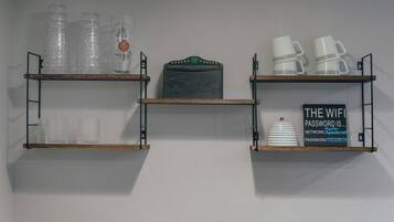 Tres repisas de madera con varios objetos: a la izquierda, botellas de plástico y un vaso con un logotipo; en el centro, un letrero con un logotipo; a la derecha, tazas blancas y un letrero con la contraseña de WiFi.