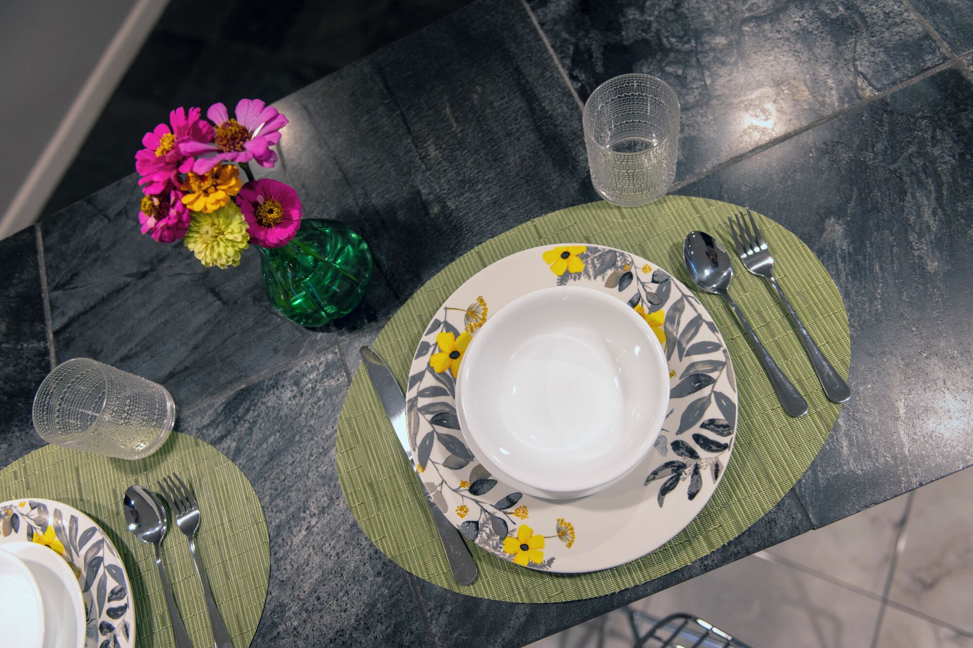 Une table dressée avec une assiette blanche, un bol, une nappe verte, un verre, une fourchette et une cuillère.