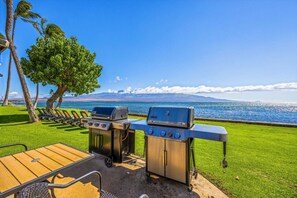 A barbecue setup with two grills, a wooden table, and chairs, overlooking a scenic ocean view. - Simply Amazing Oceanfront Condo! Milowai 310 (Wailuku)