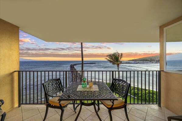 A balcony with a table and chairs, overlooking a coastal view with a sunset. - Simply Amazing Oceanfront Condo! Milowai 310 (Wailuku)