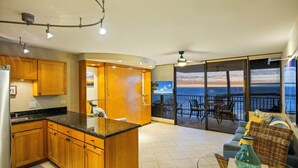 A modern kitchen with wooden cabinets and a central island, a balcony with a view of the sea, and a seating area with a wicker chair and a glass table.