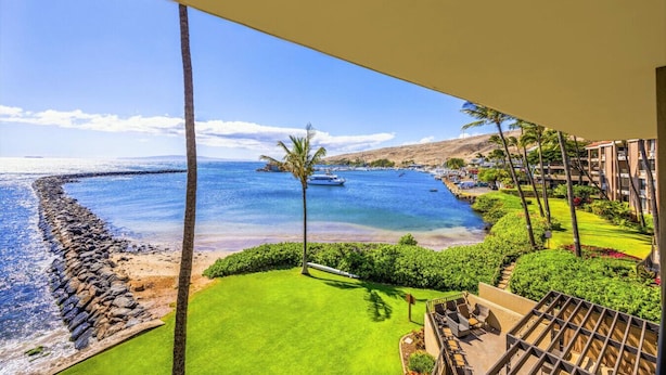 A balcony with a view of a beach, a pool, and a building with a garden.