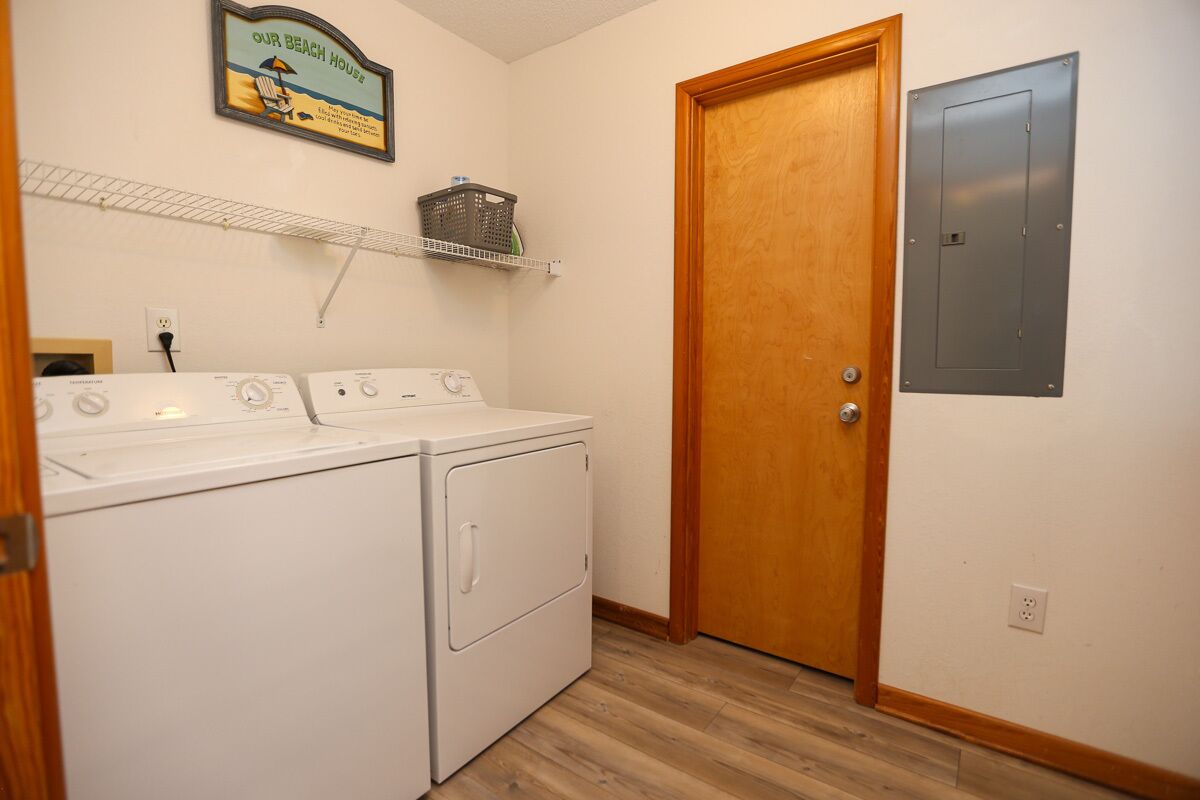A laundry room with a washer and dryer, a wooden door, a shelf with a basket, and a wall-mounted electrical box.