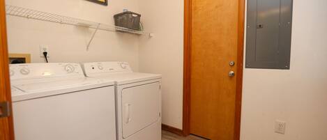 A laundry room with a washer and dryer, a wooden door, a shelf with a basket, and a wall-mounted electrical box.