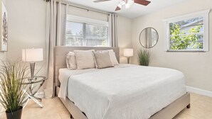 Een slaapkamer met een bed, plafondventilator, raam met zonwering, nachtkastje, lamp en een pot met een plant.