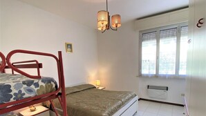 A room with a bunk bed, a single bed, a bedside table with a lamp, a window with blinds, and a radiator. - Cosy apartment for 6 guests with TV and balcony (Rosignano Marittimo)