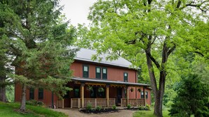 A two-story wooden house with a porch, surrounded by trees and a gravel driveway.