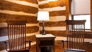 A log cabin room with wooden floors, two rocking chairs, a small table with a lamp, and a window.