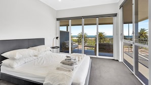 Una camera da letto moderna con un ampio letto, vista sulla spiaggia tramite delle porte scorrevoli in vetro e un balcone con mobili da esterno.