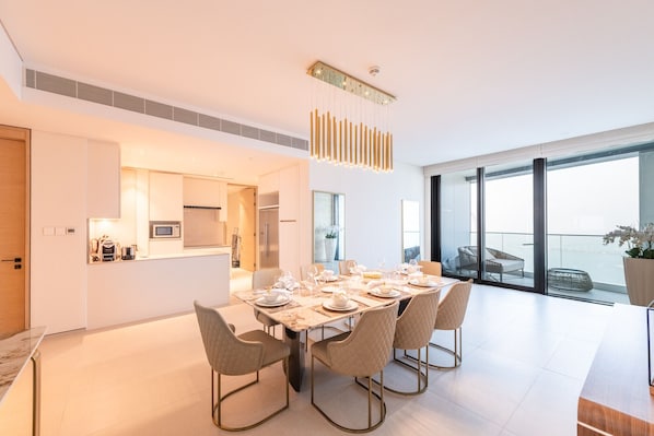 A modern dining area with a long table set for four, a kitchen in the background, and large windows offering a view of the outdoors.