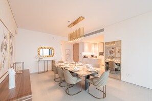 A dining area with a long table set for six, surrounded by six chairs. There is a mirror above the table and a kitchen area visible through an open door.
