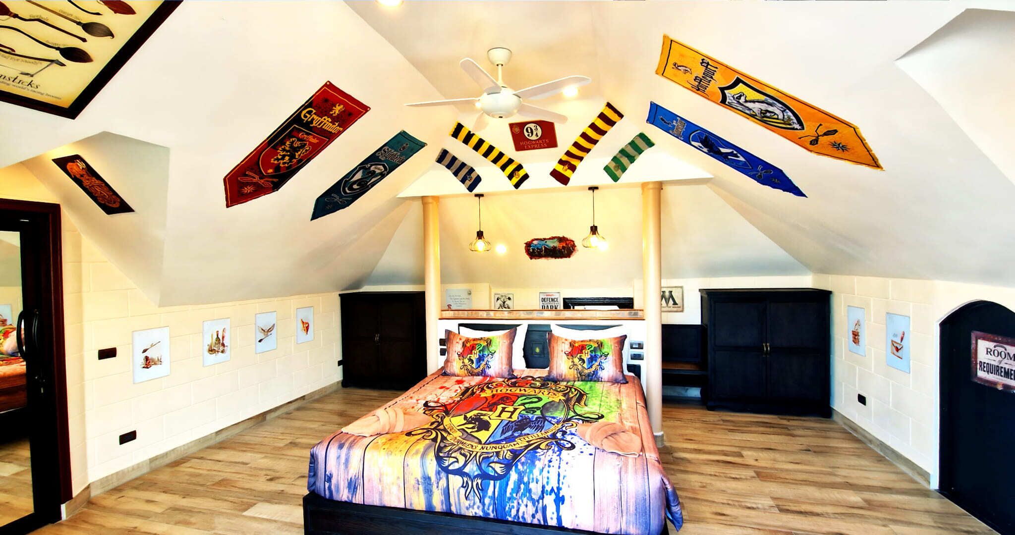 A bedroom with a sloped ceiling, a bed with a colorful Hogwarts-themed duvet, and various Hogwarts house banners hanging from the ceiling.
