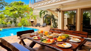 Un coin petit-déjeuner aménagé au bord de la piscine, avec une table, des chaises et une variété d’aliments pour le petit-déjeuner.
