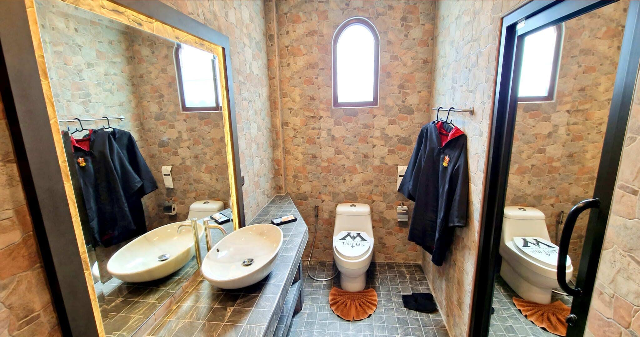 Une salle de bain avec des murs en pierre, un grand miroir, deux lavabos et deux toilettes.