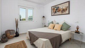 Ein Schlafzimmer mit einem Bett, Kissen, einem Gemälde, einem Deckenventilator, einem Fenster und einem kleinen Tisch mit einer Statue.