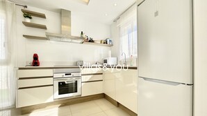 A modern kitchen with white cabinetry, a built-in oven, a refrigerator, and a window with curtains. - Allegra 19 Ciudad Quesada by Villas&You (Ciudad Quesada)