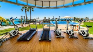 Een sportschool met loopbanden, elliptische trainers en hometrainers, met uitzicht op een zwembad en palmbomen.