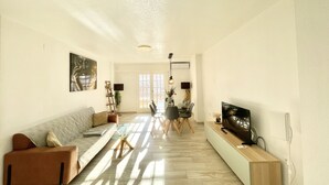 A spacious living room with a sofa, a glass coffee table, a dining area with chairs, and a TV on a stand. - Sunset Torrevieja by Villas&You (Torrevieja)