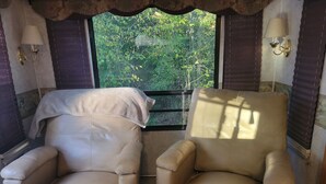 Two beige armchairs with a view of greenery through a window with blinds.