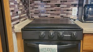 A kitchen with a black stove, a tiled backsplash, and a 'Welcome to our Happy Place' towel on the oven.
