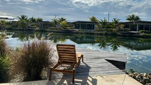 Una terraza de madera con un banco da a una masa de agua tranquila, rodeada de vegetación tropical y edificios modernos.