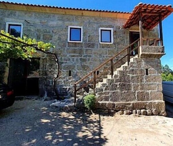 A stone building with a staircase leading to a door, surrounded by vegetation.