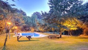Zona de piscina con una mesa y bancos, rodeada de árboles e iluminada por luces.