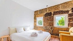 Un dormitorio con pared de piedra, cama con ropa de cama blanca, suelo de madera y dos ventanas que dan a la vegetación.