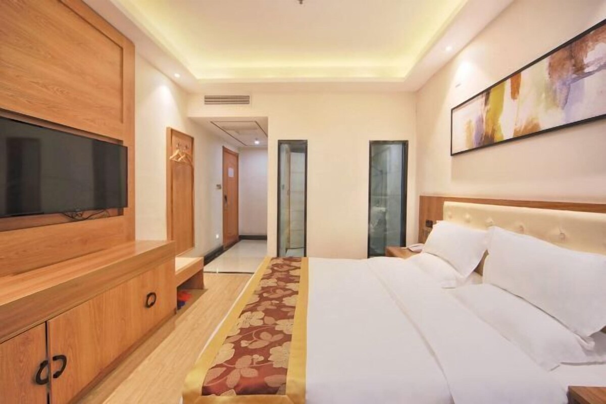 A hotel room with a large flat-screen TV, a bed with white linens, a wooden cabinet, and a painting on the wall.